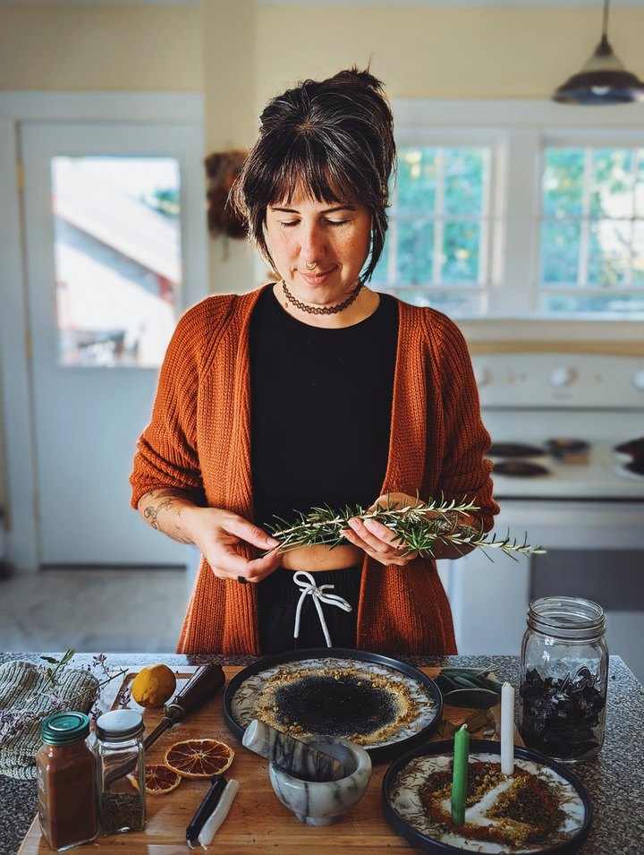 Mai in her kitchen holding a sprig of rosemary over a table of herbs, candles and dried orange slices